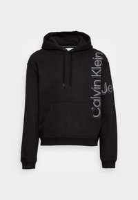 Sudadera con capucha negra, con bolsillo frontal y logo vertical "Calvin Klein Jeans" en gris en el hombro izquierdo y la manga.