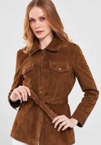 Bruine suède jas met kraag, knoopsluiting, twee borstzakken en een ceintuur in de taille. Heeft een gladde textuur en een getailleerde pasvorm.