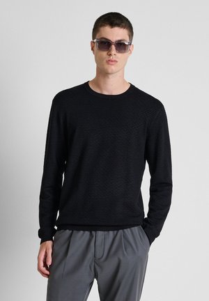 Jonge man met donkere zonnebril, zwart textuur langemouwen shirt en grijze broek, staand tegen een effen lichte achtergrond.
