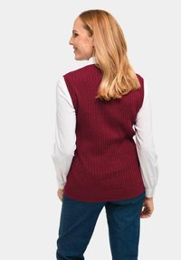 Bordeaux gebreide vest met geribbelde textuur en mouwloos ontwerp, gedragen over een wit longsleeve shirt, gecombineerd met blauwe spijkerbroeken.