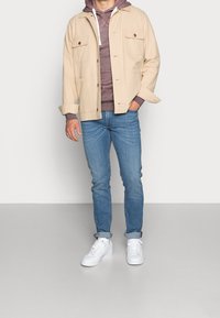 Hombre vistiendo una chaqueta beige sobre una sudadera color burdeos, jeans azules arremangados en los tobillos y zapatillas blancas, de pie contra un fondo blanco liso.