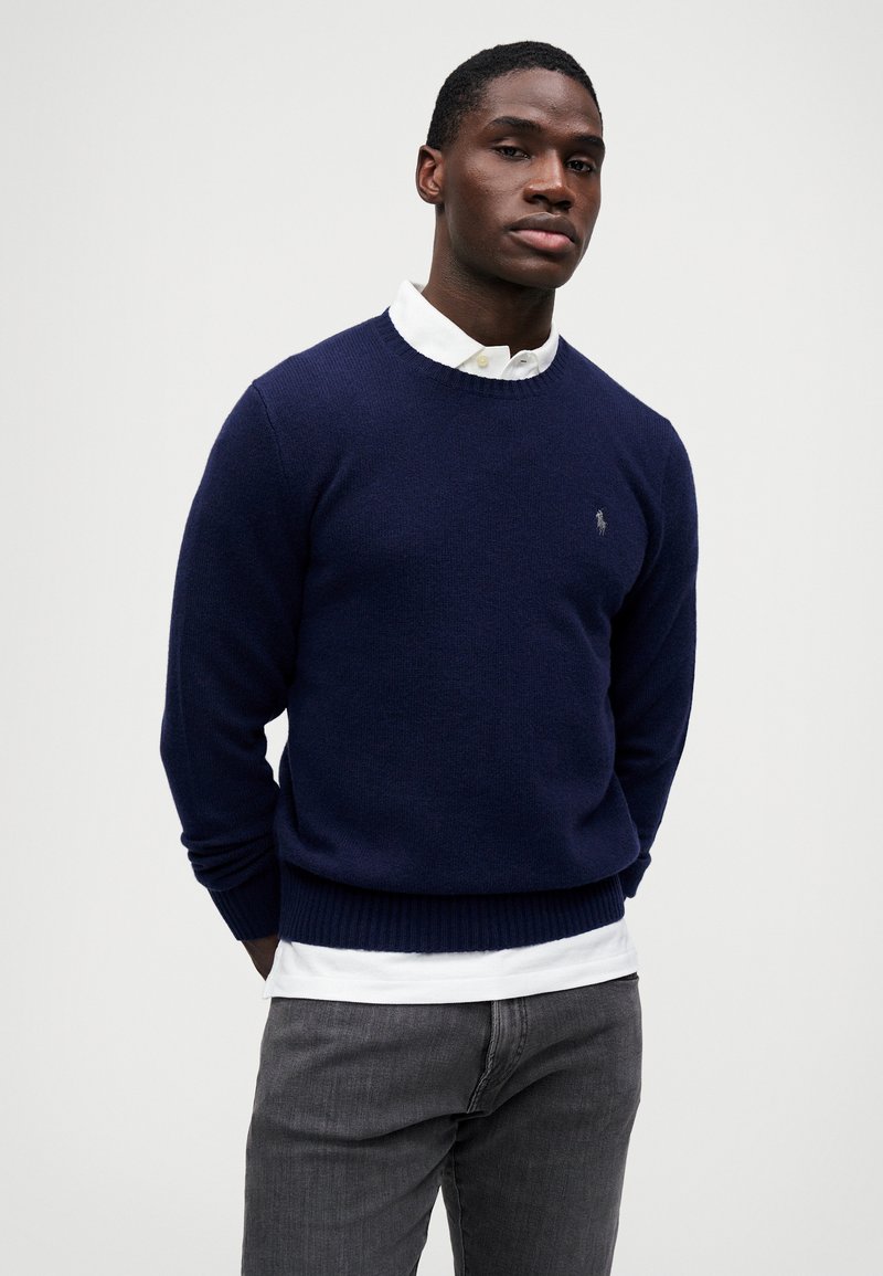 Marineblauwe crewneck trui van breiwerk, met ribgebreide manchetten en onderrand. Gelaagd over een witte blouse met kraag, gecombineerd met grijze jeans.