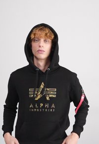 Felpa nera con cappuccio, caratterizzata da un logo grafico camouflage sul petto, cappuccio con cordoncino, tasca a marsupio e una tasca con zip con dettaglio rosso.