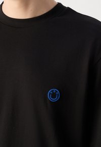 T-shirt noire avec un petit patch brodé bleu représentant un visage souriant et les lettres H, G, O sur la poitrine.