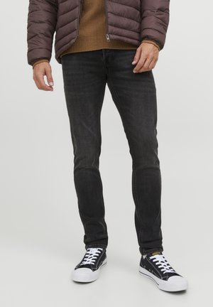 Mand iført sort slim-fit jeans, sort-hvide sneakers, brun sweater og mørkebrun quiltet jakke, stående på en ensfarvet baggrund.