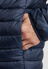 Giacca isolante blu navy con design trapuntato, che mostra una mano infilata in una tasca laterale, e un dettaglio con zip all'apertura della tasca.