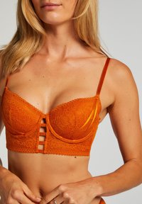 Valkoinen pitsinen bralette kirkkaan oranssina, jossa on vaijerituet, säädettävät hihnat ja kolme hakasta koostuva suljin vaakasuorilla raoilla edessä.