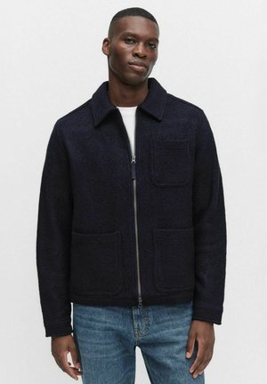 Marinblå ulljacka med zip-stängning, två framfickor och krage. Avslappnad passform med texturerad tyg. Kombinerad med ljusblå jeans.