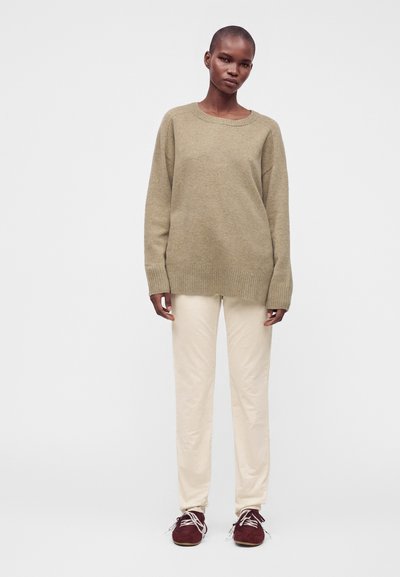 Femme portant un pull oversize beige, un pantalon crème et des baskets bordeaux, debout devant un fond blanc uni.