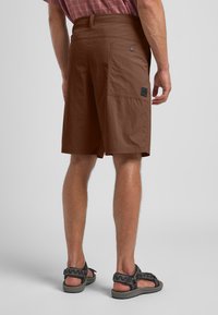 Bruine cargoshorts met een gladde textuur, zijzak en een zwart labeldetail. Gecombineerd met donkere sandalen met een gepatternde band.