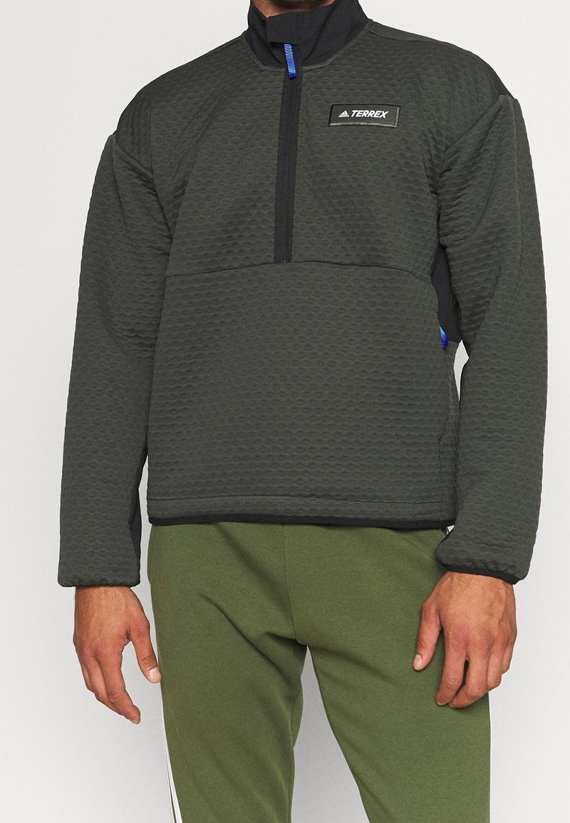 Homme portant un pull texturé gris foncé avec une demi-fermeture éclair et le logo Terrex sur la poitrine, associé à un pantalon vert olive.