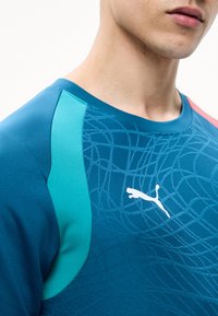 Άνδρας που φορά μπλε αθλητική μπλούζα Puma με αφηρημένο σχέδιο και ανοιχτό μπλε πάνελς στους ώμους, περικομμένη στη λαιμό και το άνω μέρος του κορμού.