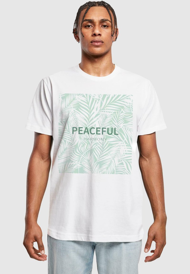 White cotton t-shirt featuring a green palm leaf graphic and the word "PEACEFUL" above "HARMONY" in a bold font. Casual design, relaxed fit.