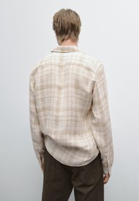 Camicia a righe beige con bottoni e maniche lunghe, caratterizzata da una texture morbida e un orlo curvo. Indossata con pantaloni scuri, vista da dietro.