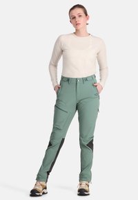 Hellgrüne Outdoor-Hosen mit schwarzen Akzenten, mehreren Taschen und einer taillierten Passform, kombiniert mit einem cremefarbenen Langarmoberteil und Wanderschuhen.