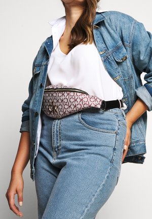 Vrouw met een spijkerjack en spijkerbroek, wit shirt, met een patroonrijke CK-riemtasje rond de taille.