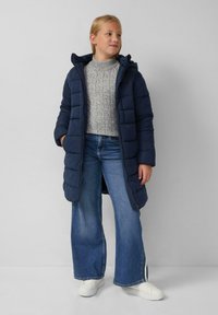 Marineblauwe pufferjas met een capuchon, met een gevoerd ontwerp. Gecombineerd met een grijze kabeltrui en wijde blauwe jeans. Witte sneakers maken de look af.