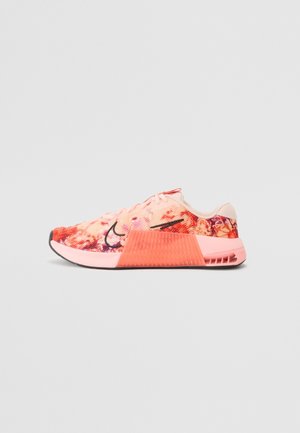 Roze en oranje sportschoen met bloemenpatroon, zwart swoosh-logo en dikke, getextureerde tussenzool, zijaanzicht op een effen achtergrond.