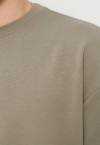 Κοντινή λήψη ενός ατόμου που φοράει απλό, ανοιχτό καφέ μπλουζάκι με στρογγυλή λαιμόκοψη και ορατές ραφές κοντά στον γιακά και τον ώμο.