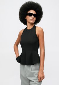 Žena s veľkým afrom, v čiernej bezrukávovej peplum blúzke, svetlosivých prúžkovaných nohaviciach a veľkých čiernych slnečných okuliaroch, pózujúca s jednou rukou v vrecku.