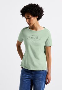 Chemise à manches courtes vert clair avec un col rond, présentant un design textuel réfléchissant sur le devant. Assortie à un jean bleu.