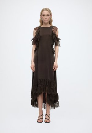 Žena stojí oblečená v čiernych bezrukávových šatách s čipkovými volánmi na ramenách a leme, skombinovaných s čiernymi sandálmi so šnúrkami na jednoduchom pozadí.