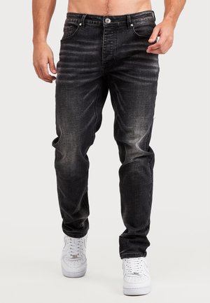 Mann trägt ausgewaschene schwarze Jeans und weiße Sneakers, dargestellt von der Taille bis zu den Füßen vor einem schlichten Hintergrund.