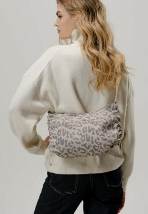 Vrouw met golvend blond haar, gekleed in een witte trui en zwarte broek, met een beige schoudertas met luipaardprint over haar rug gedragen.