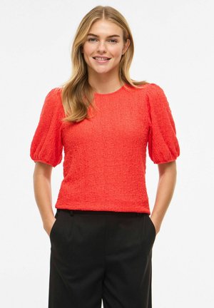 Mladá žena sa usmieva, má na sebe červenú textúrovanú blúzku s nadýchanými rukávmi a čierne nohavice, stojí s rukami v vreckách na bielom pozadí.