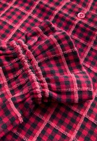 Sarkans un melns šallīgs flaneļa audums ar mīkstu tekstūru un izteiktām rūtiņām. Iespējotas rozā pogas un cieši piegulošs piedurknes dizains.