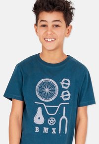 T-paita puuvillasta teal-värissä, jossa on valkoiset BMX-pyöräkuviot, mukaan lukien rengas, polkimet, satula ja ohjaustanko. Lyhyet hihat ja pyöreä pääntie.