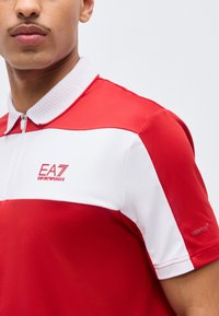Κοντινό πλάνο ατόμου που φοράει κόκκινο και λευκό πόλο με φερμουάρ, με το λογότυπο EA7 Emporio Armani στο στήθος και τη σήμανση Ventus στο μανίκι.
