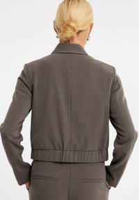Brun cropped blazer med en tekstureret overflade, klapkrave og samlet kant. Har lange ærmer og diskret sømdetaljering.