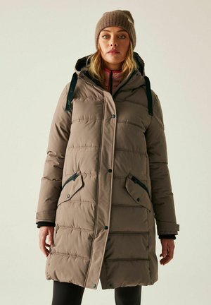 Kobieta ubrana w beżową pikowaną zimową kurtkę z kapturem i czarnymi kieszeniami na zamek błyskawiczny, w zestawieniu z brązową dzianą czapką i czarnymi legginsami.