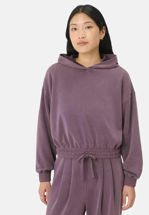 Vrouw draagt een losse paarse hoodie met lange mouwen, elastische taille en bijpassende paarse joggingbroek met trekkoord tegen een effen achtergrond.