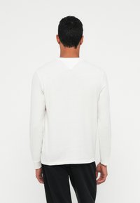 Homem com cabelo preto curto, vestindo uma camisa branca de mangas longas simples e calças pretas, de pé, de costas, contra um fundo branco.