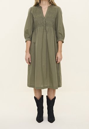 Olivengrøn bomuldskjole med V-hals, smocket buste og trekvart lange pufærmer. Kombineret med sorte ruskinds cowboystøvler.