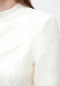 Witte stof met pailletten en een gestructureerd oppervlak, voorzien van een hoge halslijn. De mouwen zijn glad en aangesloten, wat een gestructureerde uitstraling geeft.