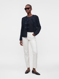 Femme portant une veste noire texturée avec des nœuds, un haut noir, un pantalon blanc, des chaussures plates noires et des lunettes de soleil, debout, une main dans la poche.