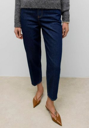 Personne portant un jean court bleu marine, un pull gris et des chaussures à talons aiguilles beige à lanière, se tenant sur un sol de couleur claire.