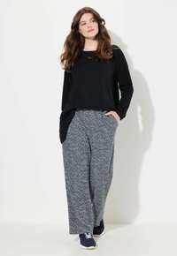 Vrouw met lang bruin haar, gekleed in een zwarte lange mouwtop, blauwgrijze wijde broek en marineblauwe sneakers, staand tegen een witte achtergrond.