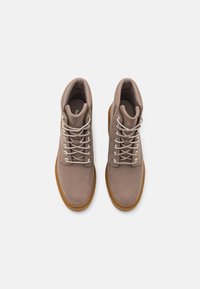 Bottes chevilles en daim marron avec bouts ronds, œillets en métal et lacets gris. Les semelles sont en caoutchouc avec une couleur gomme contrastante.