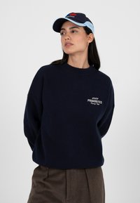 Femme portant un pull bleu marine "Prohibited Racing Club", une casquette bicolore bleu marine et bleu clair, et un pantalon marron, regardant sur le côté contre un fond gris.