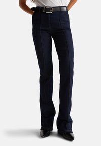 Tmavě modré zvonové džíny s kontrastním švem, vysokým pasem a černým opaskem s obdélníkovou sponou. Hladká denimová textura.