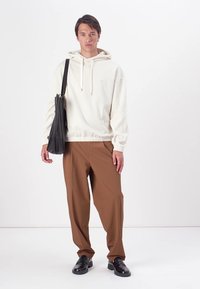 Lichtbeige hoodie met trekkoorden, losse pasvorm, geribde zoom; bruine wijde broek; zwarte schoenen; zwarte schoudertas.