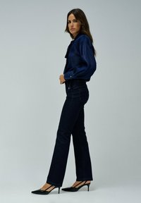 Jeans de mezclilla oscuros acampanados combinados con una blusa azul marino con cuello y detalles de botones, complementados con tacones negros de punta.