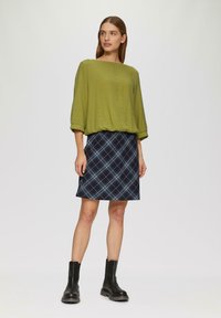Groene textuur top met wijde halslijn en driekwart mouwen, gecombineerd met een marineblauwe geruite rok. Zwarte enkelboots maken de look af.