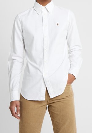 Personne portant une chemise blanche à boutons avec un petit logo brodé sur la poitrine et un pantalon en velours côtelé marron clair, main dans la poche.
