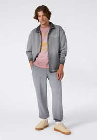 Grijze jas met een zwarte kraag boven een roze graphic t-shirt, gecombineerd met grijze sweatpants en beige sneakers, en een relaxte pasvorm.