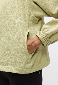 Veste vert clair texturée avec poignets élastiqués et poches latérales. Présente un logo discrètement brodé sur le devant.